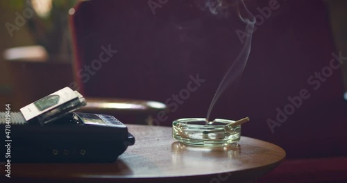 Low angle home interior detail shot of side table with vintage tape recorder / answering machine and ashtray that has a lit CBD marijuana pre-roll joint, cigarette with smoke rising