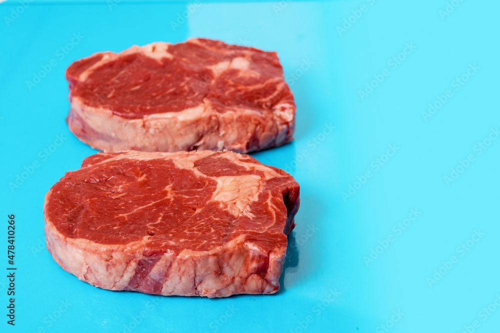 Two fresh juicy rib eye steak on blue surface. Top quality fresh meat. Butcher craft product and meat industry. Rich red color and marbling. Premium cut. Two fresh juicy rib eye steak on blue surface. Top quality fresh meat. Butcher craft product and meat industry. Rich red color and marbling. Premium cut.
