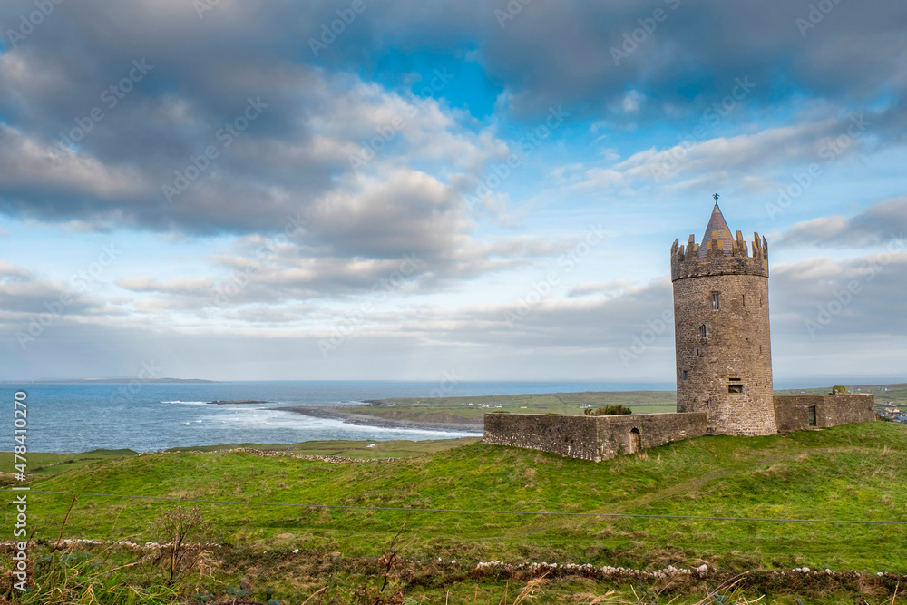 Doonagore Castle on a green hill, Doolin pier and Atlantic ocean in the background. Beautiful cloudy sky. Popular tourist attraction. History preservations. Amazing old stronghold and fort.