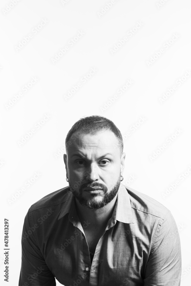 Obraz premium Emotive portrait of bearded charismatic muscular 40 years old man sitting over white background, looking at camera. Short haircut. Black and white studio shot. Text space