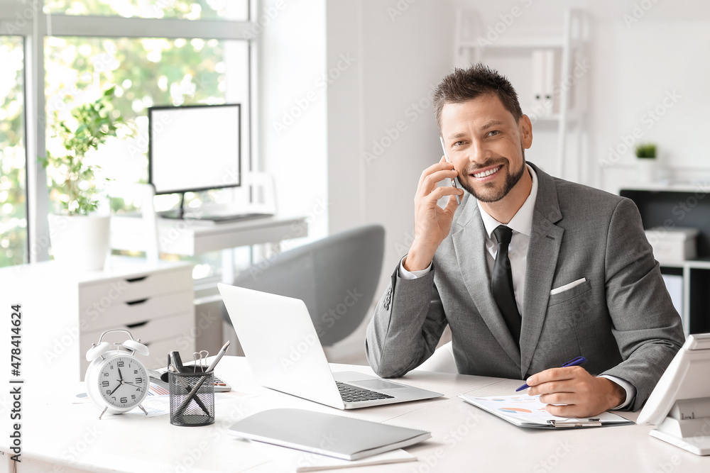 Smiling businessman talking by mobile phone at table in office Smiling businessman talking by mobile phone at table in office