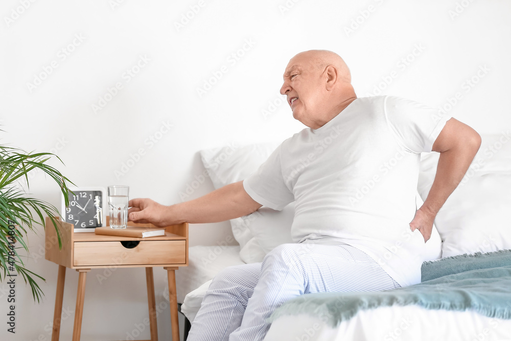Obraz premium Senior man with back pain taking glass of water from table in bedroom