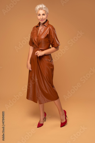 Beautiful blonde middle aged woman with short hairstyle posing in brown medium dress. Fashion photo. Elegant businesswoman. Full photo