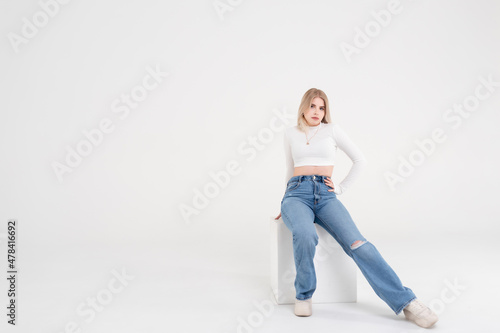 Young blonde girl in a white sweater and jeans sits and poses on an isolated white background in the studio. People lifestyle concept. Copy space for copy