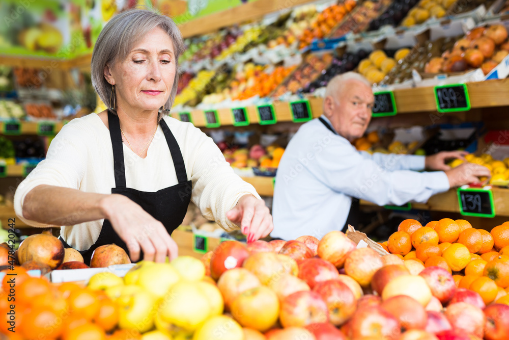 Woman merchandiser in apron putting goods on shelf in greengrocer