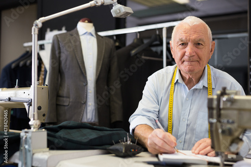 Elderly man tailor uses notepad to record jacket measurements in workshop