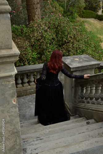  portrait of pretty  female model with red hair wearing glamorous gothic black lace ballgown.  Posing in a fairytale castle location with staircases 