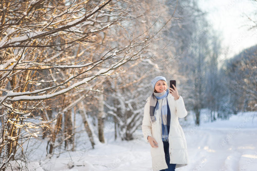 A beautiful blonde woman in a white winter coat and a blue hat walks through a snow-covered park on a winter day and takes pictures on a smartphone and takes a selfie. Winter walks.