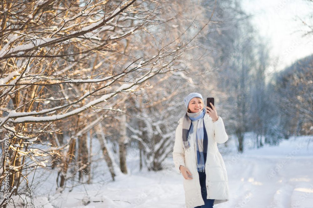 A beautiful blonde woman in a white winter coat and a blue hat walks through a snow-covered park on a winter day and takes pictures on a smartphone and takes a selfie. Winter walks. A beautiful blonde woman in a white winter coat and a blue hat walks through a snow-covered park on a winter day and takes pictures on a smartphone and takes a selfie. Winter walks.