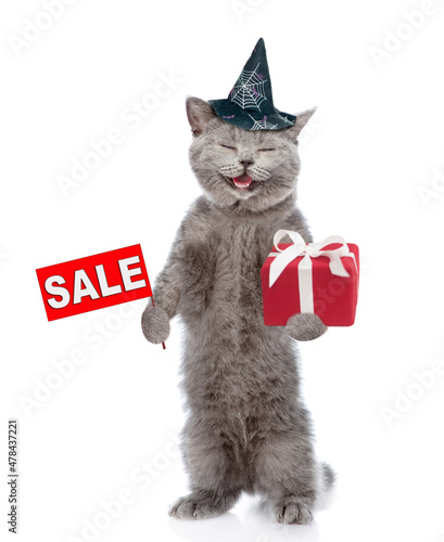 Happy cat wearing hat for halloween stands on hind legs holds gift box and shows sales symbol . isolated on white background