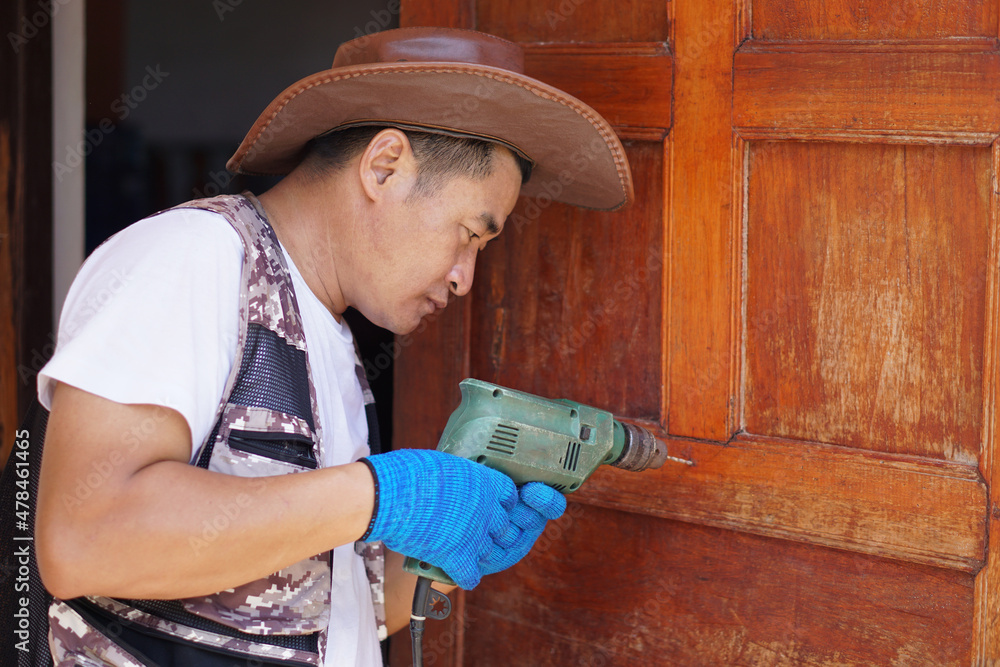 Handsome Asian male carpenter is using electric drill to fix wooden door. Concept : Home renovation. Repair or fixing. Construction, handyman, tool and repair in daily life. DIY work. Handsome Asian male carpenter is using electric drill to fix wooden door. Concept : Home renovation. Repair or fixing. Construction, handyman, tool and repair in daily life. DIY work.