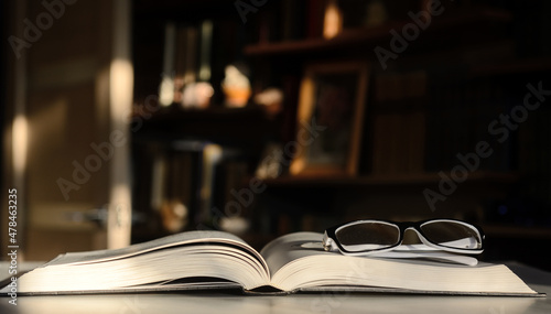 There is an open book on the table. There are glasses on the pages. The background is bookshelves illuminated by the sun.