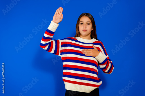 Cute Hispanic girl wearing striped sweater against blue wall. Swearing with the hand on the chest and the open palm, taking an oath of allegiance