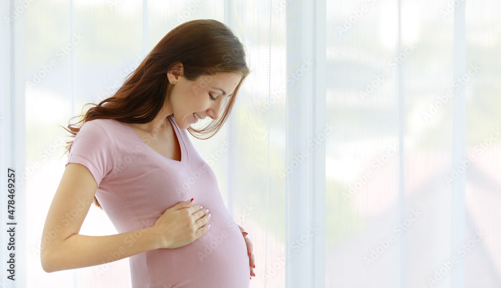 Caucasian young healthy female big belly tummy prenatal pregnant mother model in casual shirt standing in front of curtain background smiling holding glass of drinking milk in morning alone at home
