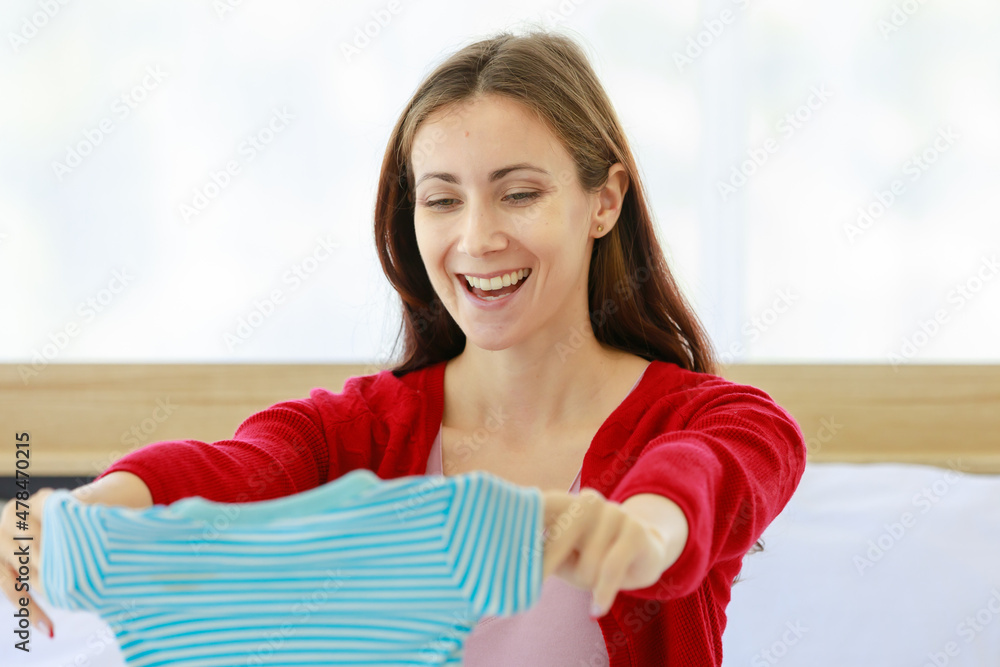 Caucasian millennial young happy female prenatal pregnant mother in casual pregnancy outfit jacket sitting smiling on bed in bedroom holding preparing choosing small cotton baby clothes in hands Caucasian millennial young happy female prenatal pregnant mother in casual pregnancy outfit jacket sitting smiling on bed in bedroom holding preparing choosing small cotton baby clothes in hands