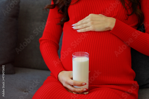 Closeup unrecognizable unknown Caucasian millennial young happy sexy female prenatal pregnant mother in casual red pregnancy dress sitting on cozy sofa smiling touching belly holding fresh milk glass