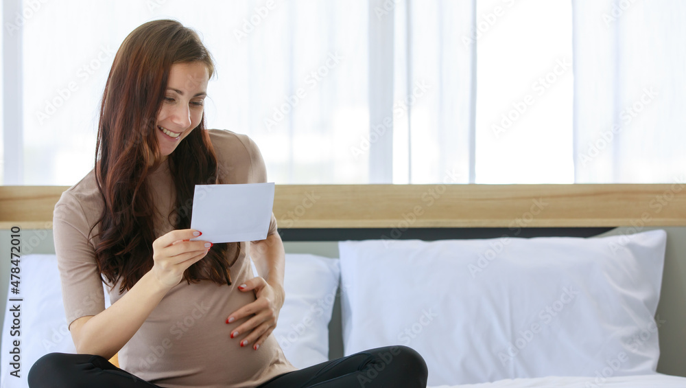 Caucasian millennial young happy female prenatal pregnant mother in casual brown pregnancy sitting on bed smiling holding showing ultrasound Xray scan picture in bedroom at home Caucasian millennial young happy female prenatal pregnant mother in casual brown pregnancy sitting on bed smiling holding showing ultrasound Xray scan picture in bedroom at home