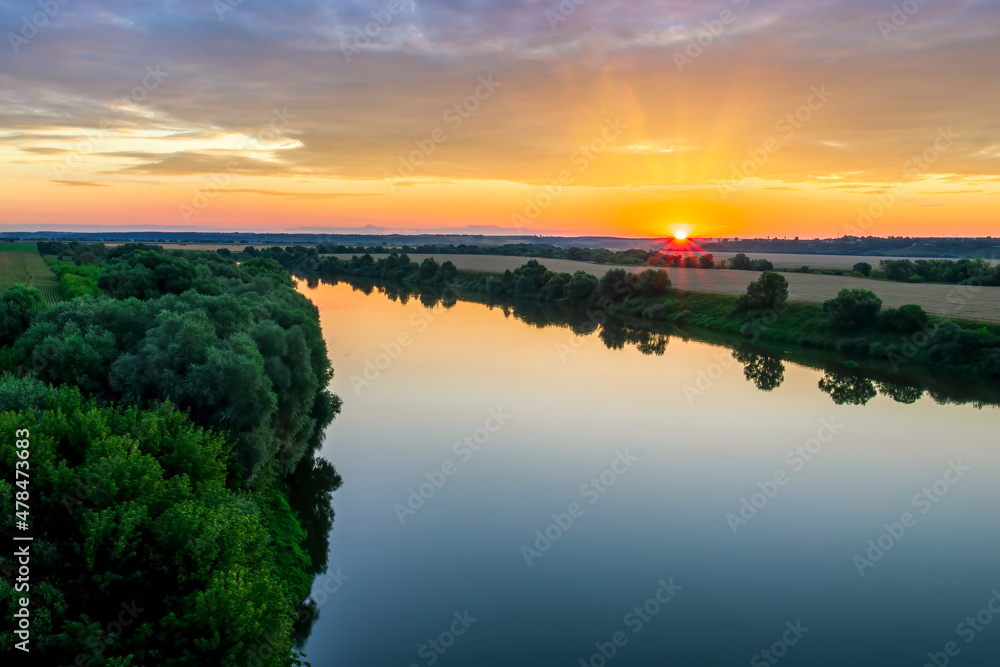 Scenic view at beautiful sunset or sunrise on a shiny river with green bushes on sides, golden sun rays, calm water ,deep blue cloudy sky and forest on a background, spring landscape