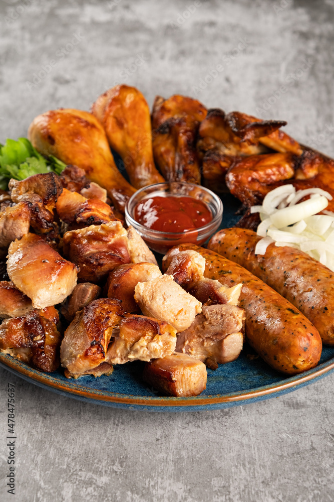 Assorted grilled meat in a plate on the table. Fried chicken legs and wings, sausages, shish kebab, pickled onions and tomato sauce. Vertical orientation, close-up, no people