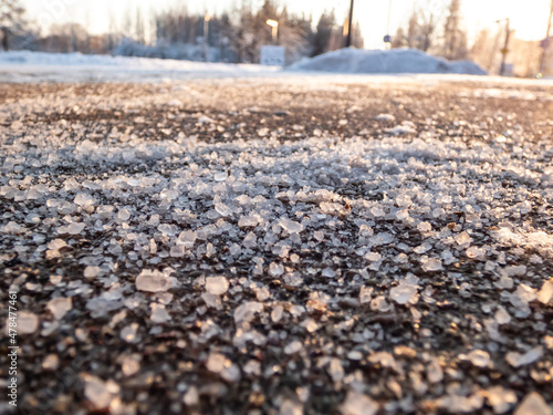 Salt grains on icy sidewalk surface in the winter. Applying salt to keep roads clear and people safe in winter weather from ice or snow. Macro view of salt grains with winter scenery in bacground