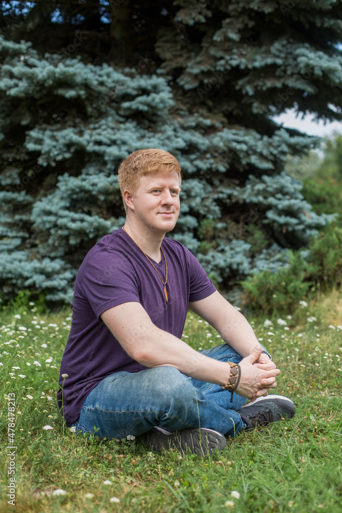 Portrait of redhead caucasian man wearing purple t-shirt and blue jeans and sitting on the grass in city park during lunch break at summer. Person looks away. Theme of rest in work break. Portrait of redhead caucasian man wearing purple t-shirt and blue jeans and sitting on the grass in city park during lunch break at summer. Person looks away. Theme of rest in work break.