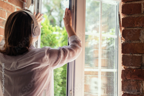 Young beautiful happy woman in headphones listening music opens window of country house and breathes fresh air in the early summer morning. Back view.