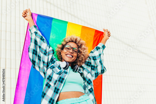 Black woman with eyeglasses, headsets and afro hair holds up the lgbtq flag of gay pride that moves with the wind on a sunny day. She fights for sexual freedom. photo