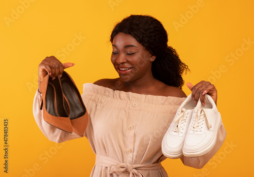 Young African American female selecting between high-heeled shoes and comfortable sneakers on orange background