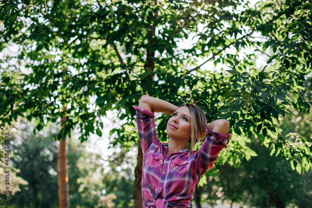 Portrait photo of adult shining blonde woman with face looking upward and hands behind head standing in park wearing checkered shirt tied in belly area in daytime. Adorable background full of green. Portrait photo of adult shining blonde woman with face looking upward and hands behind head standing in park wearing checkered shirt tied in belly area in daytime. Adorable background full of green.