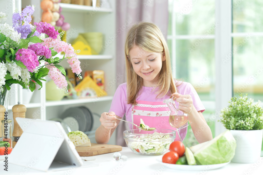 Cute little girl preparing fresh salad on kitchen table with tablet