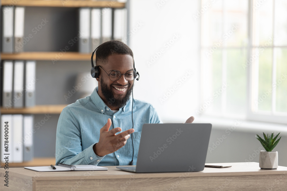Cheerful attractive millennial black male with beard in glasses, headphones gestures with hands look at pc Cheerful attractive millennial black male with beard in glasses, headphones gestures with hands look at pc