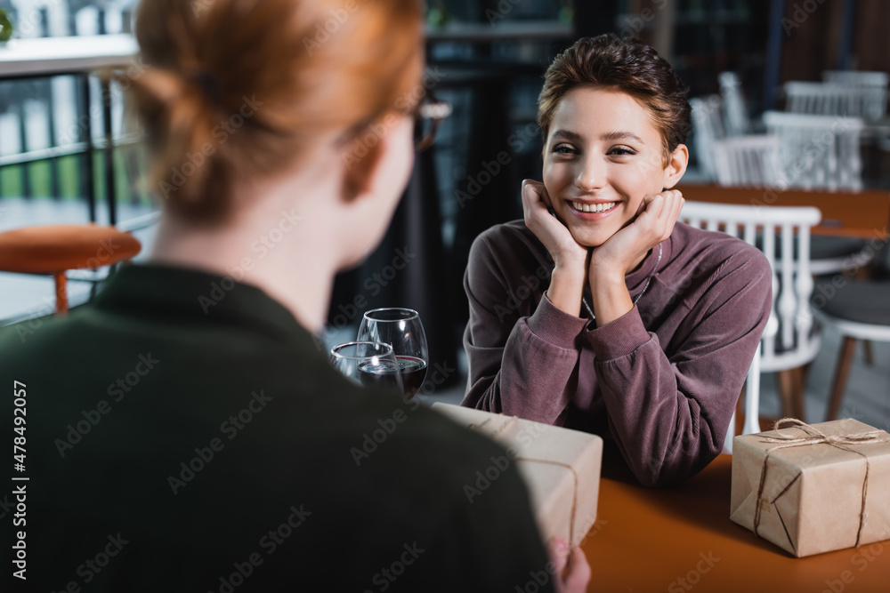 Joyful woman looking at blurred boyfriend with present near wine in hotel cafe Joyful woman looking at blurred boyfriend with present near wine in hotel cafe