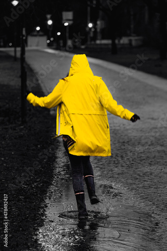 Fotografie von einer Frau, welche an einem regnerischen Tag durch die Pfützen in einem gelben, leuchtenden Regenmantel springt.