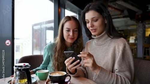 Two young attractive girls in a cafe are looking at the phone. One tells the other, teaches how to use, explains.