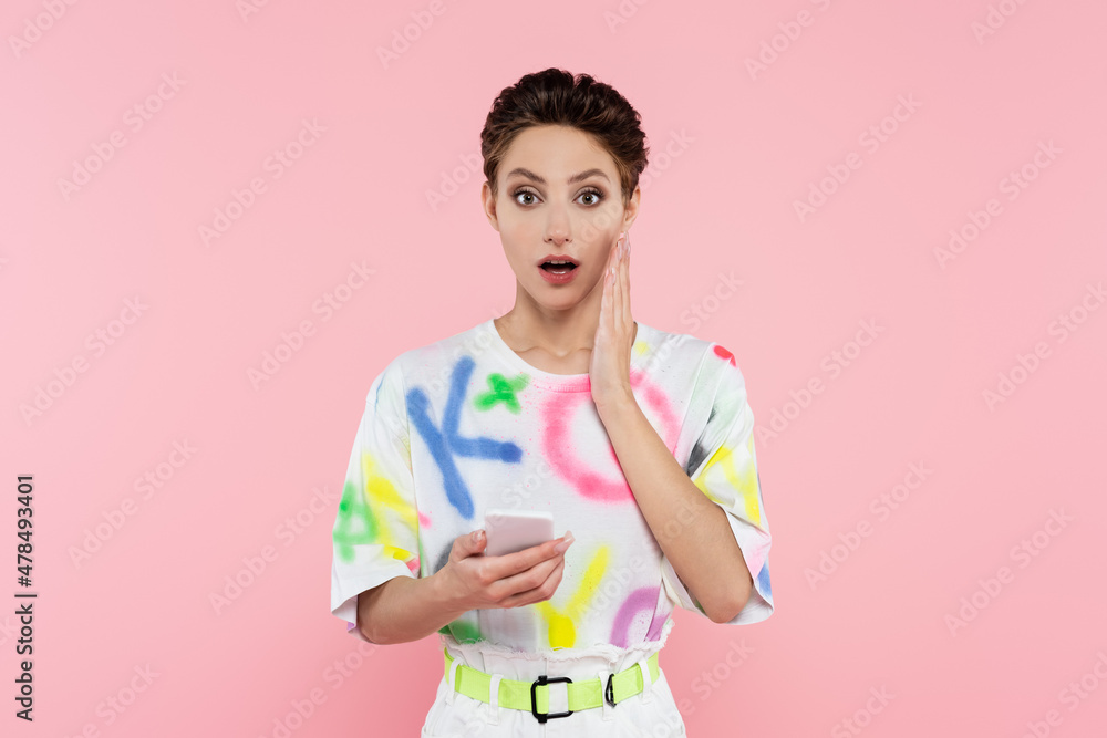 surprised woman with cellphone holding hand near face while looking at camera isolated on pink surprised woman with cellphone holding hand near face while looking at camera isolated on pink