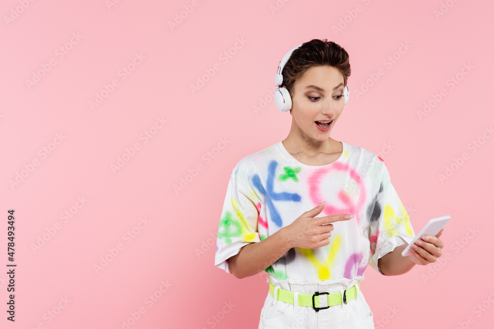amazed woman in headphones pointing at smartphone isolated on pink amazed woman in headphones pointing at smartphone isolated on pink