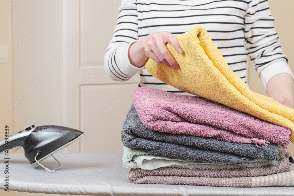 Woman stands by an ironing board with an iron and is folding a stack of  ironed,