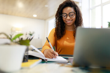 Woman sitting at desk, using computer and writing in notebook