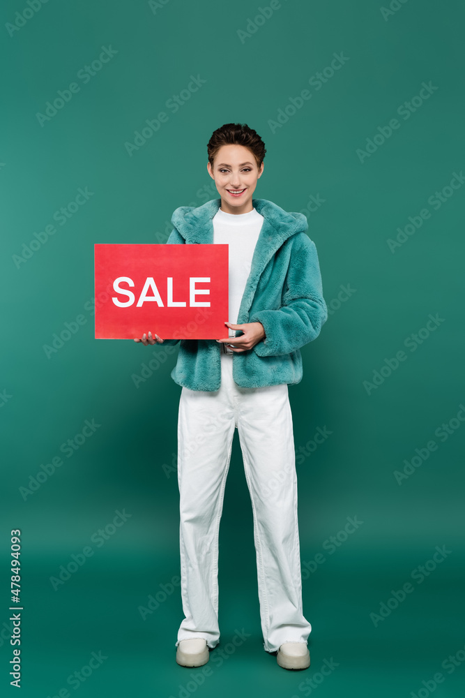 full length view of happy woman in white trousers and faux fur jacket holding red card with sale lettering on green full length view of happy woman in white trousers and faux fur jacket holding red card with sale lettering on green