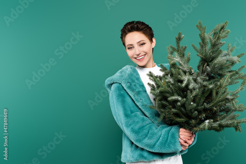 cheerful woman in trendy faux fur jacket holding small christmas tree isolated on green
