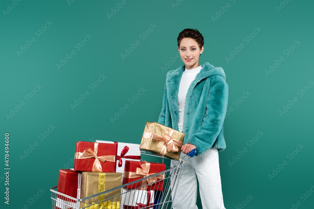 brunette woman in faux fur jacket and white trousers smiling near shopping cart with presents isolated on green brunette woman in faux fur jacket and white trousers smiling near shopping cart with presents isolated on green