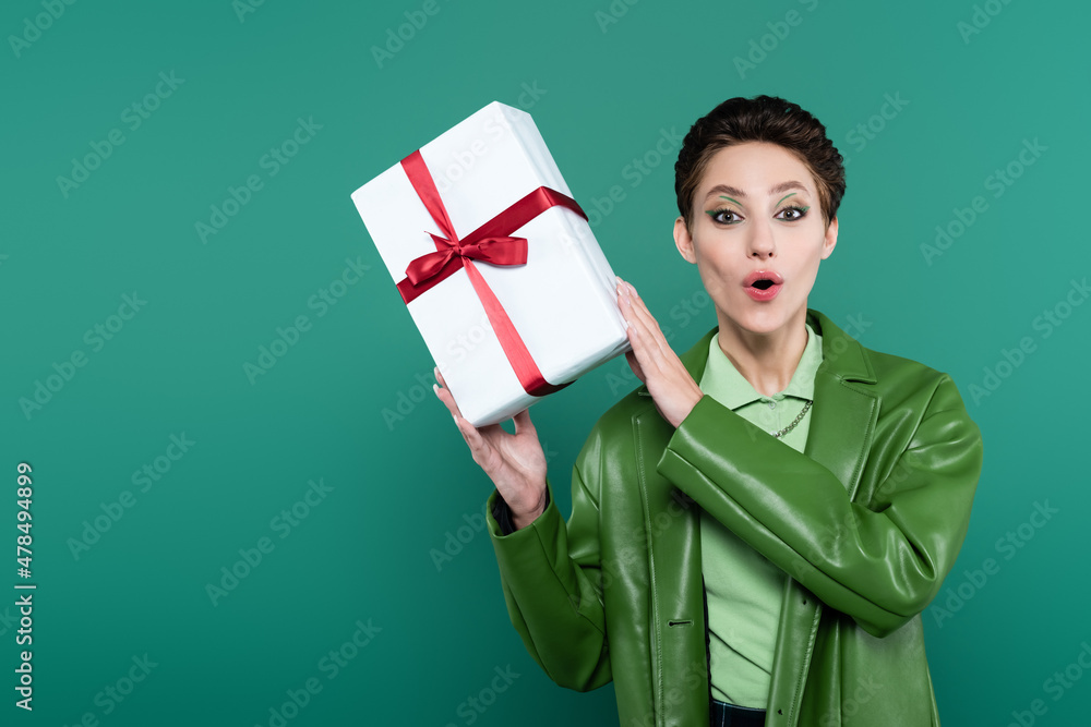 astonished woman in trendy jacket holding white gift box and looking at camera on green astonished woman in trendy jacket holding white gift box and looking at camera on green