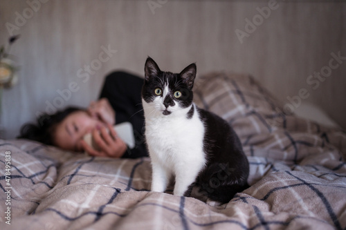 Young Asian woman using phone while petting young black and white cat on top of bed with warm sheets in bedroom