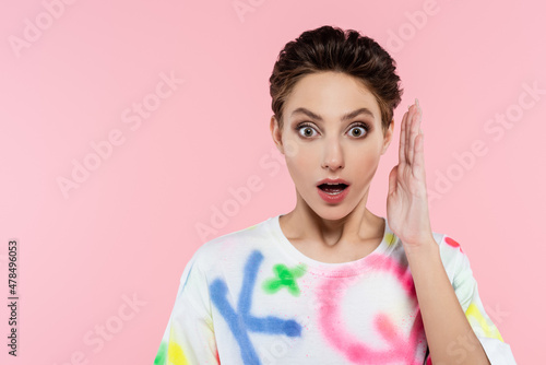 thrilled woman with open mouth holding hand near face while looking at camera isolated on pink