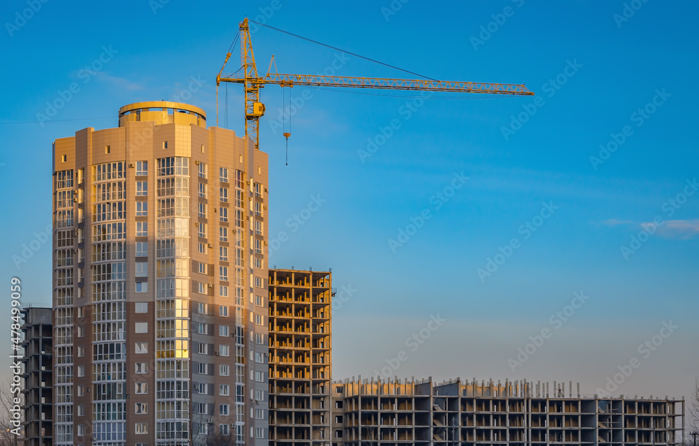 view of an unfinished non residential building with a high rise crane illuminated by the rays of the sun at sunset against a blue sky with an empty space for inserting text view of an unfinished non residential building with a high rise crane illuminated by the rays of the sun at sunset against a blue sky with an empty space for inserting text