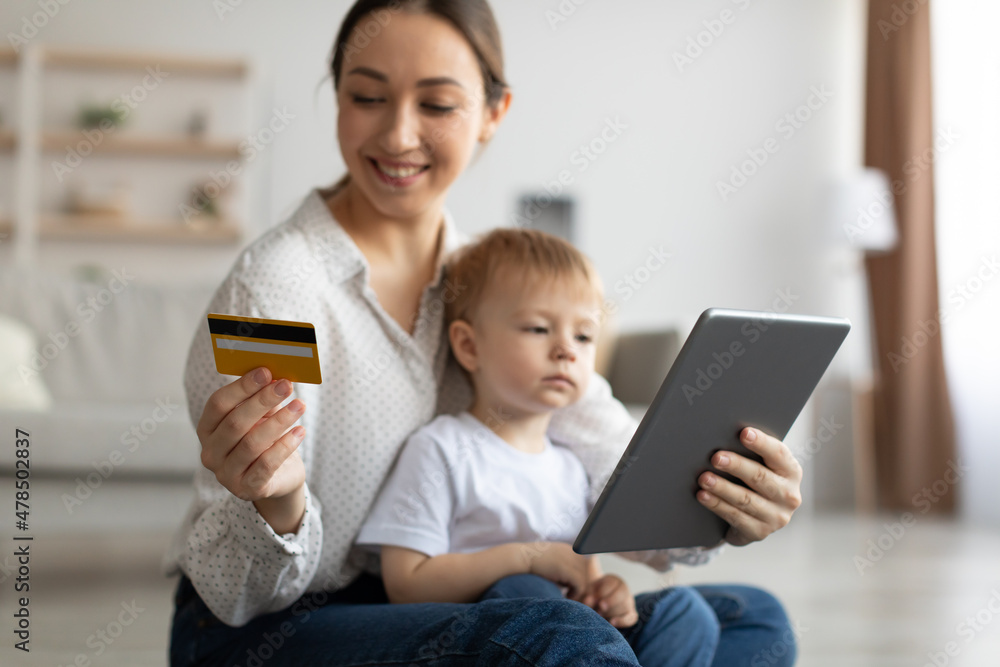Happy mother with child using digital tablet and credit card, purchasing in internet, sitting on floor in living room Happy mother with child using digital tablet and credit card, purchasing in internet, sitting on floor in living room