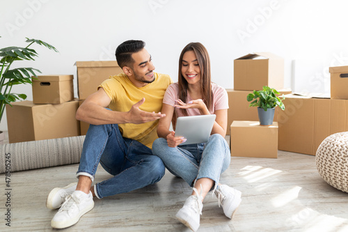 Married young diverse couple having online video call with relatives, sitting on floor among boxes on relocation day