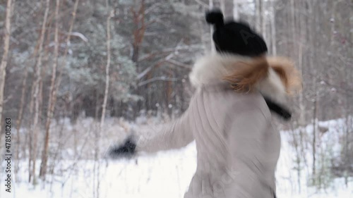 Red-haired girl throws snow to the top, smiling, and dancing in winter forest in warm clothes: black hat and light jacket, 4k. Blurred forest background