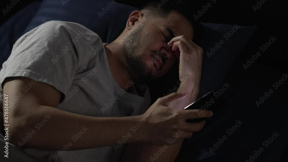 Young Asian man having sore and tired eyes when using smartphone while lying in bed at night. Young man cannot sleep from insomnia Young Asian man having sore and tired eyes when using smartphone while lying in bed at night. Young man cannot sleep from insomnia