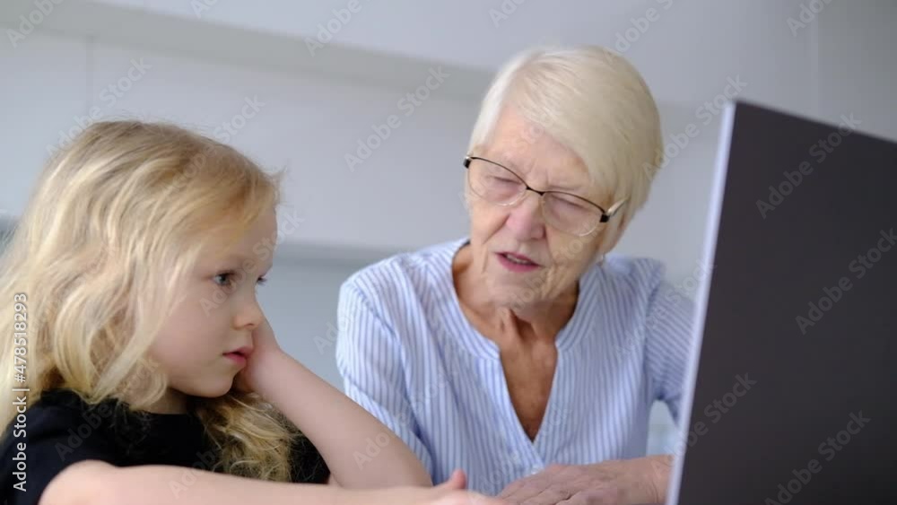 elderly caucasian woman grandmother and little curly granddaughter using laptop in the kitchen at home. online learning concept. woman with child spend time together elderly caucasian woman grandmother and little curly granddaughter using laptop in the kitchen at home. online learning concept. woman with child spend time together